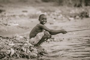 a grayscale photo of a boy sitting near the river while holding a wooden stick