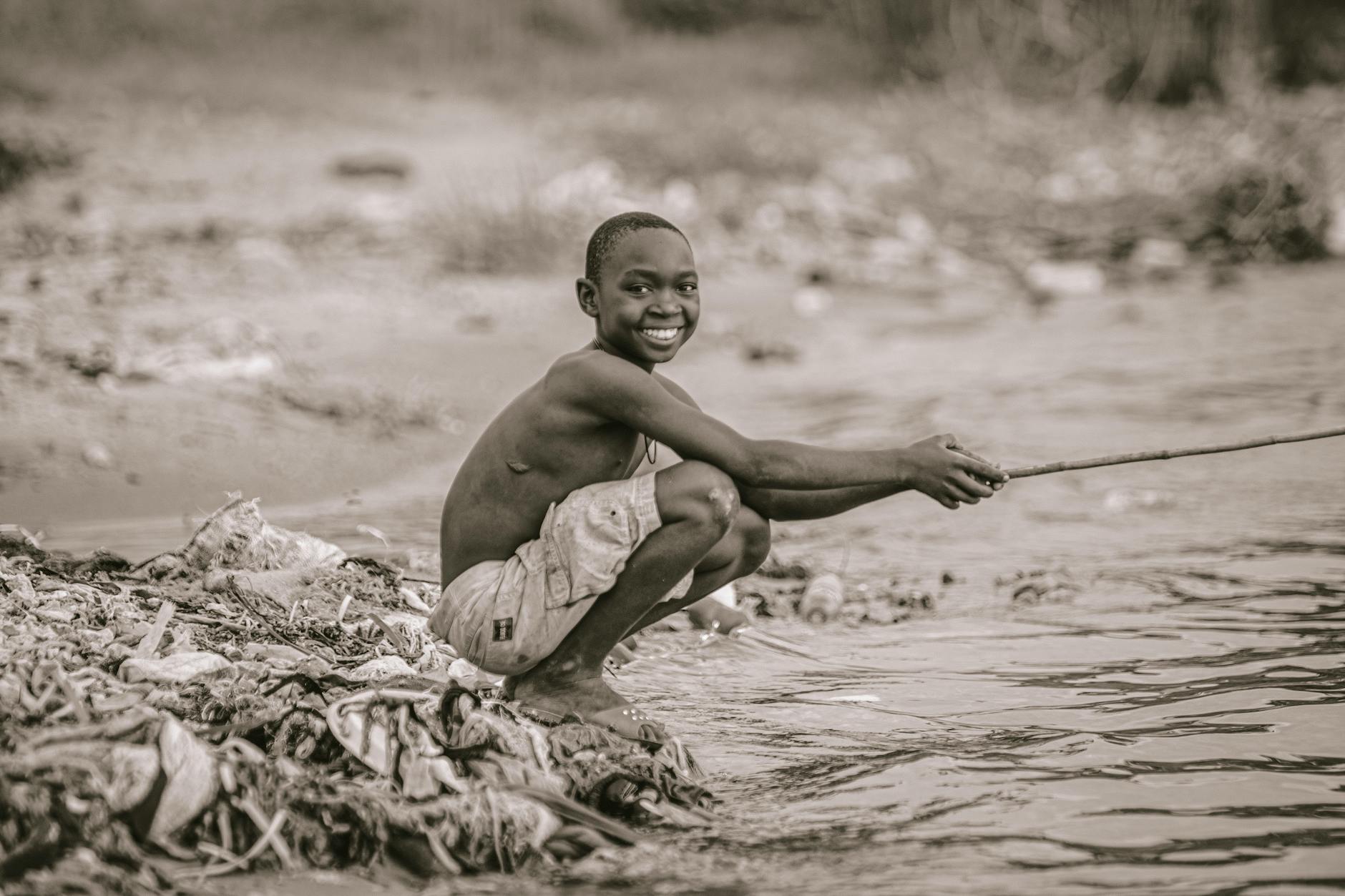 a grayscale photo of a boy sitting near the river while holding a wooden stick
