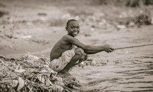 a grayscale photo of a boy sitting near the river while holding a wooden stick